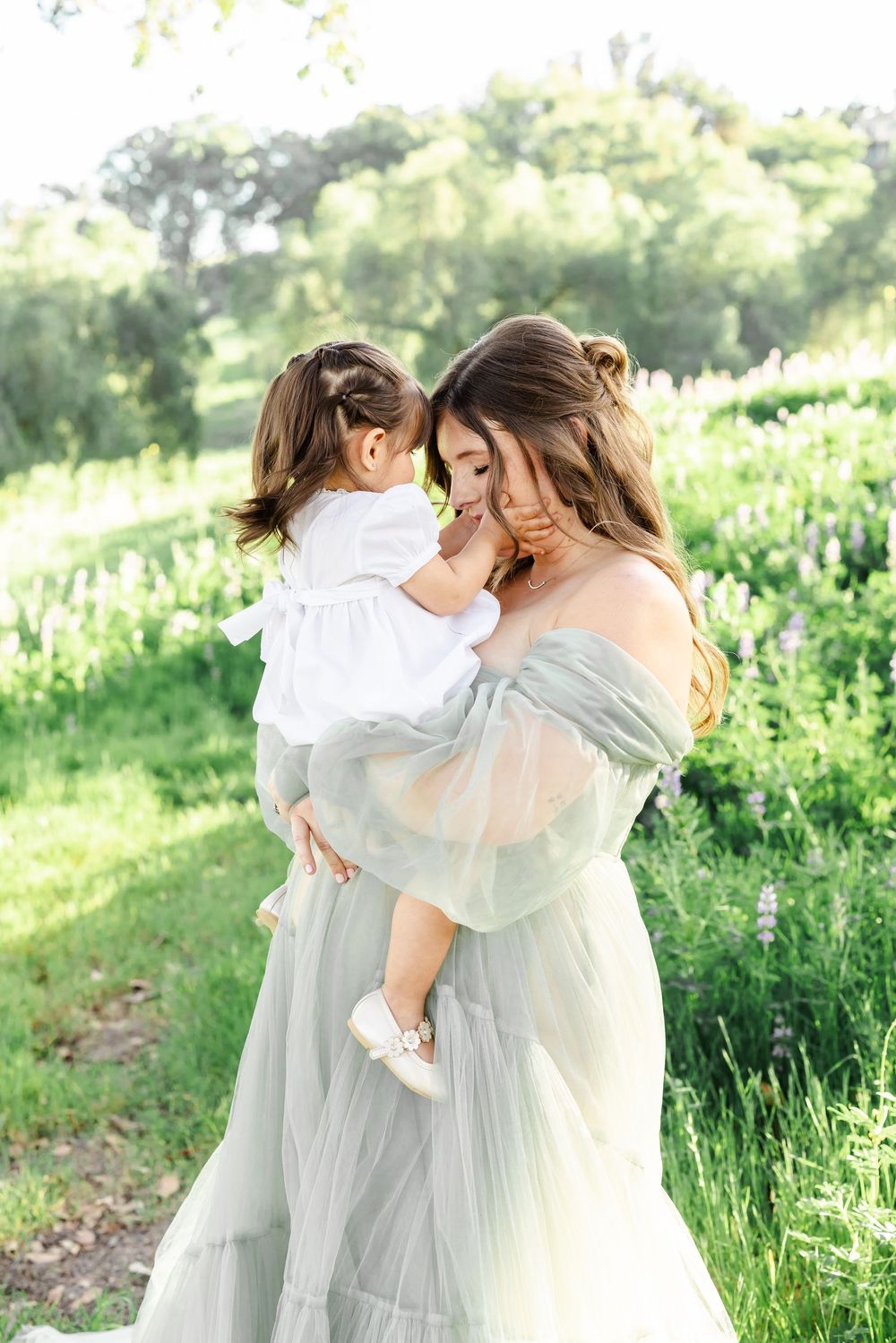 A mother in a sage green tulle gown holds her toddler in white dress, sharing a tender moment in a lush garden.