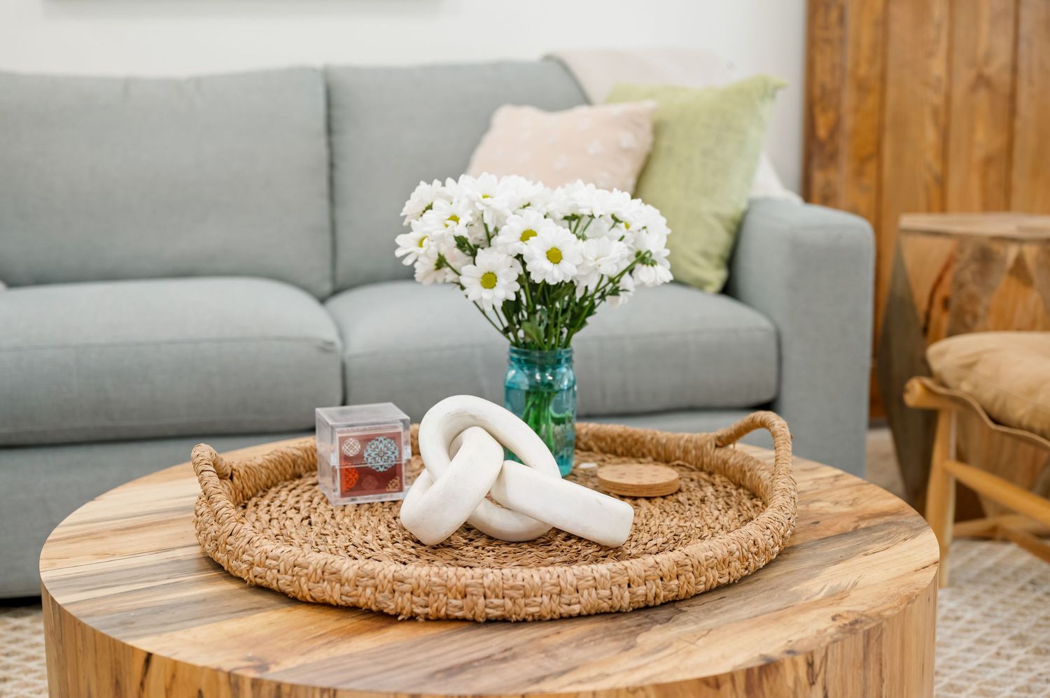 White daisies in teal vase with woven rattan tray on round wooden coffee table beside gray sofa in living room.