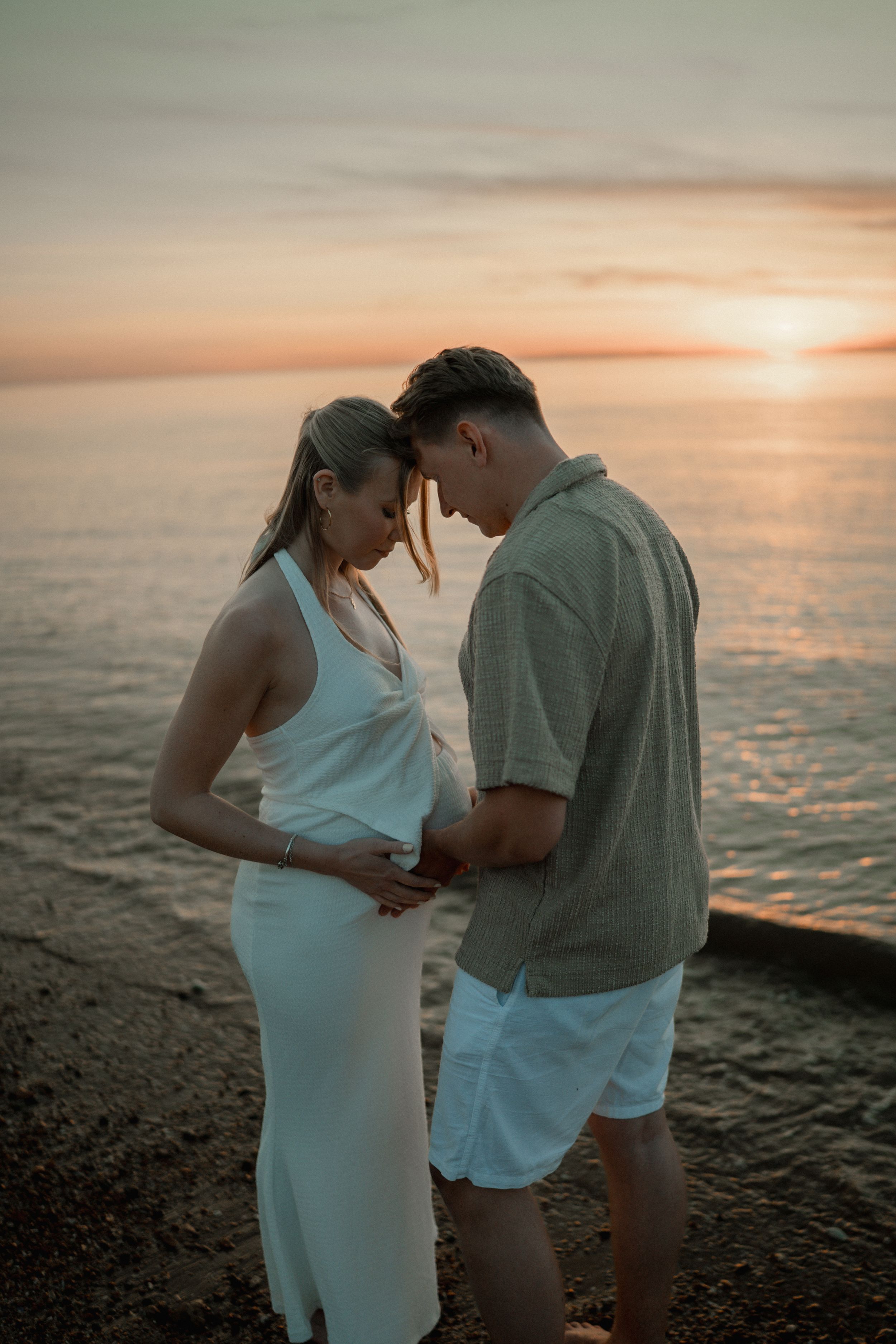 A couple in white attire shares an intimate moment on a rocky shore during golden sunset over the ocean.