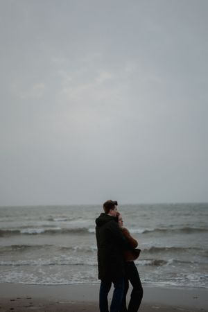 Two figures embrace while standing on a moody beach with gray skies and rolling waves in the background.