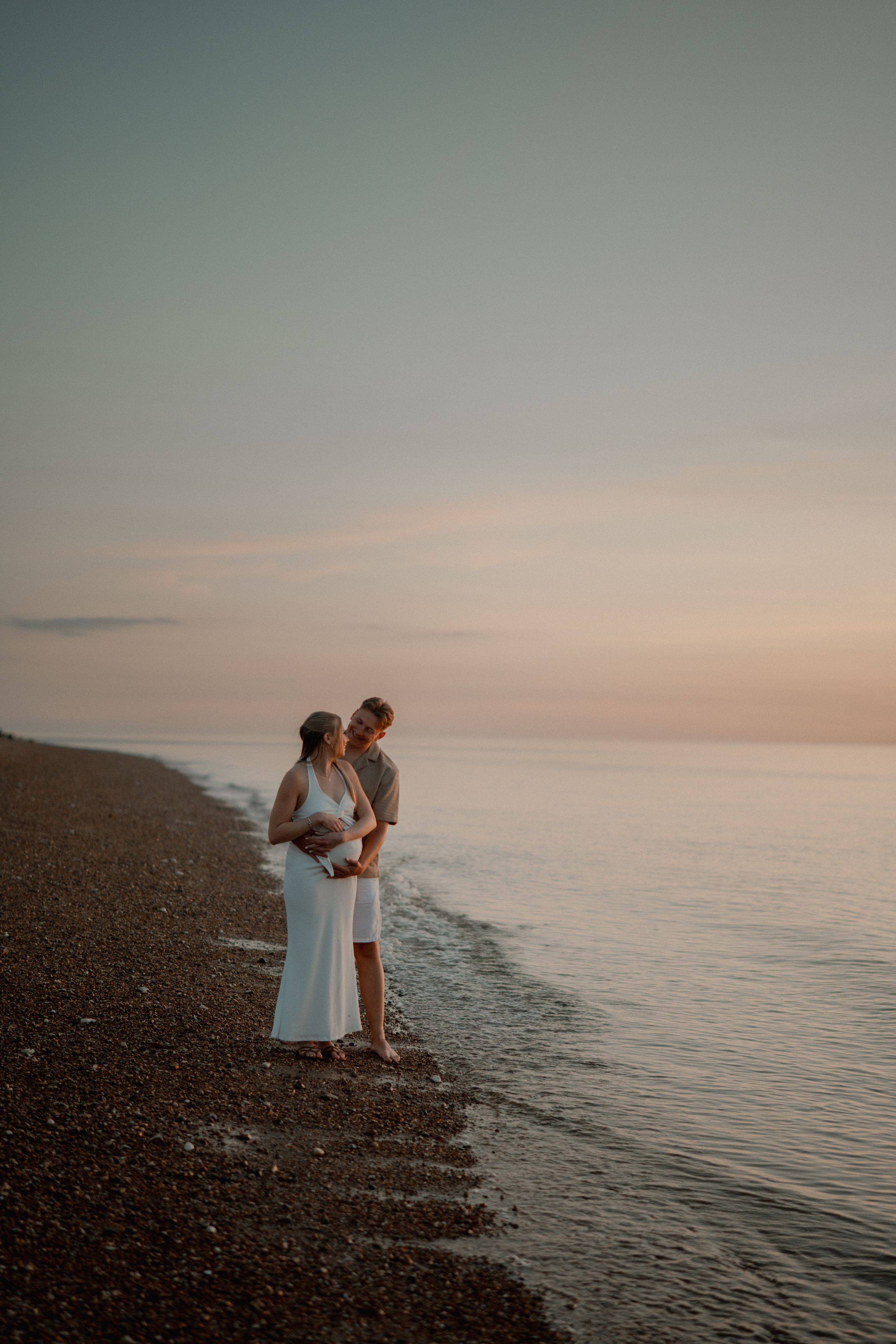 A couple stands embracing on a rocky beach at sunset with calm ocean waters and soft pink sky in the background.