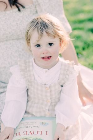 Young child in white blouse and vest holds a book while sitting outdoors on a sunny day.