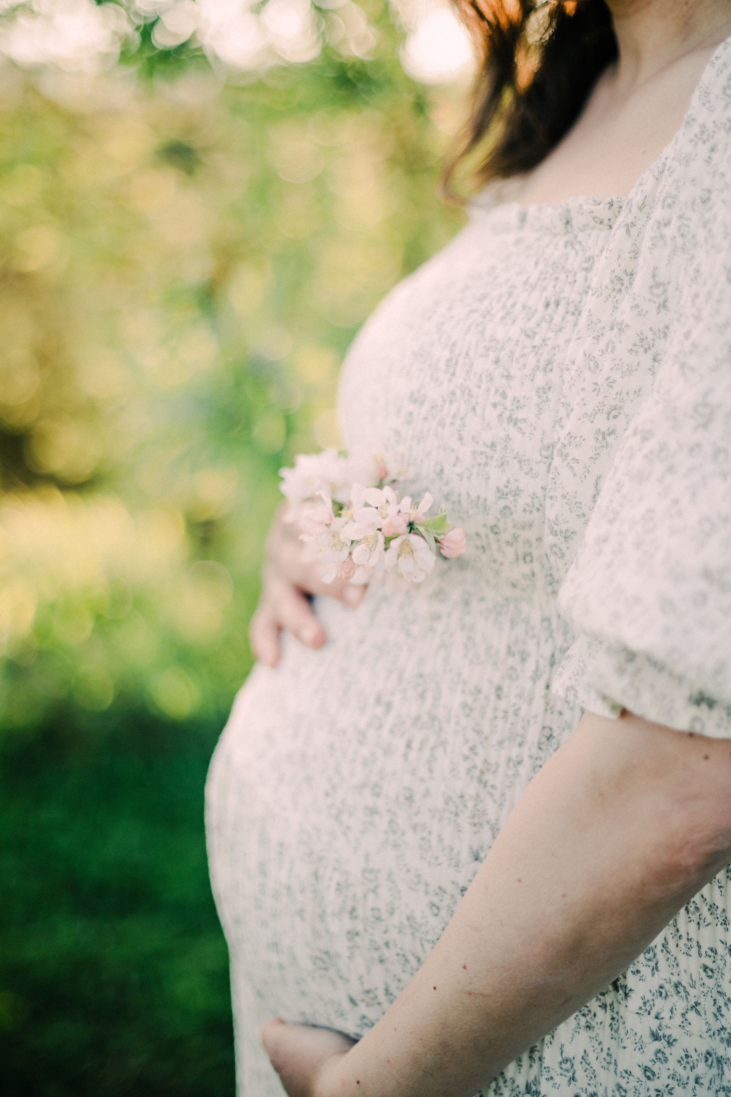 Close-up of pregnant belly in white lace dress with delicate spring flower detail against blurred outdoor background.