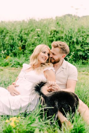 A couple sits together in grass with a black dog during an outdoor maternity photoshoot.