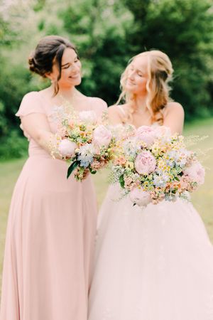 Two bridesmaids in pink dresses lean towards each other while holding delicate pastel floral bouquets in an outdoor setting.