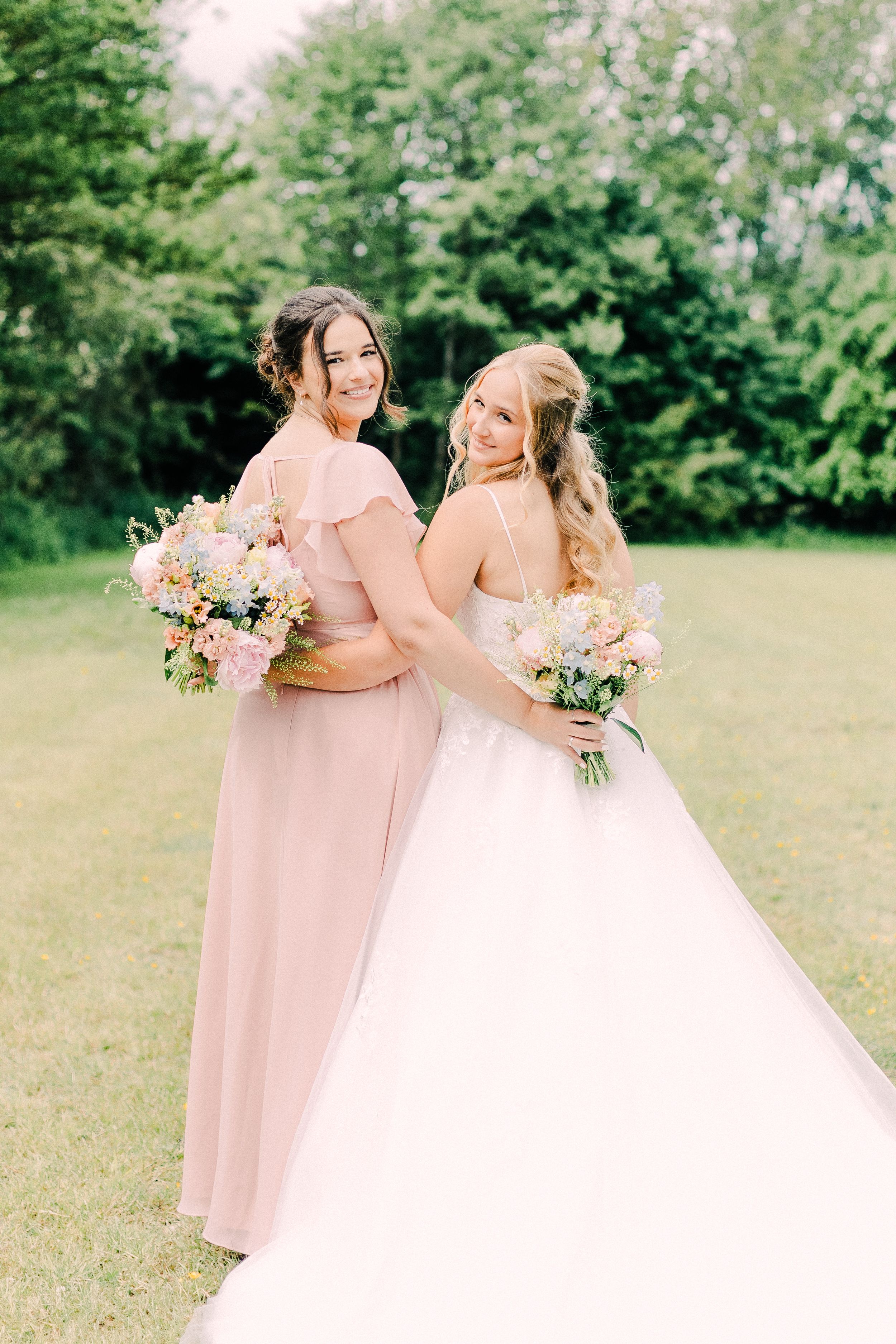 A row of bridesmaids in blush pink dresses walk elegantly with the bride in a white gown through a lush garden setting.
