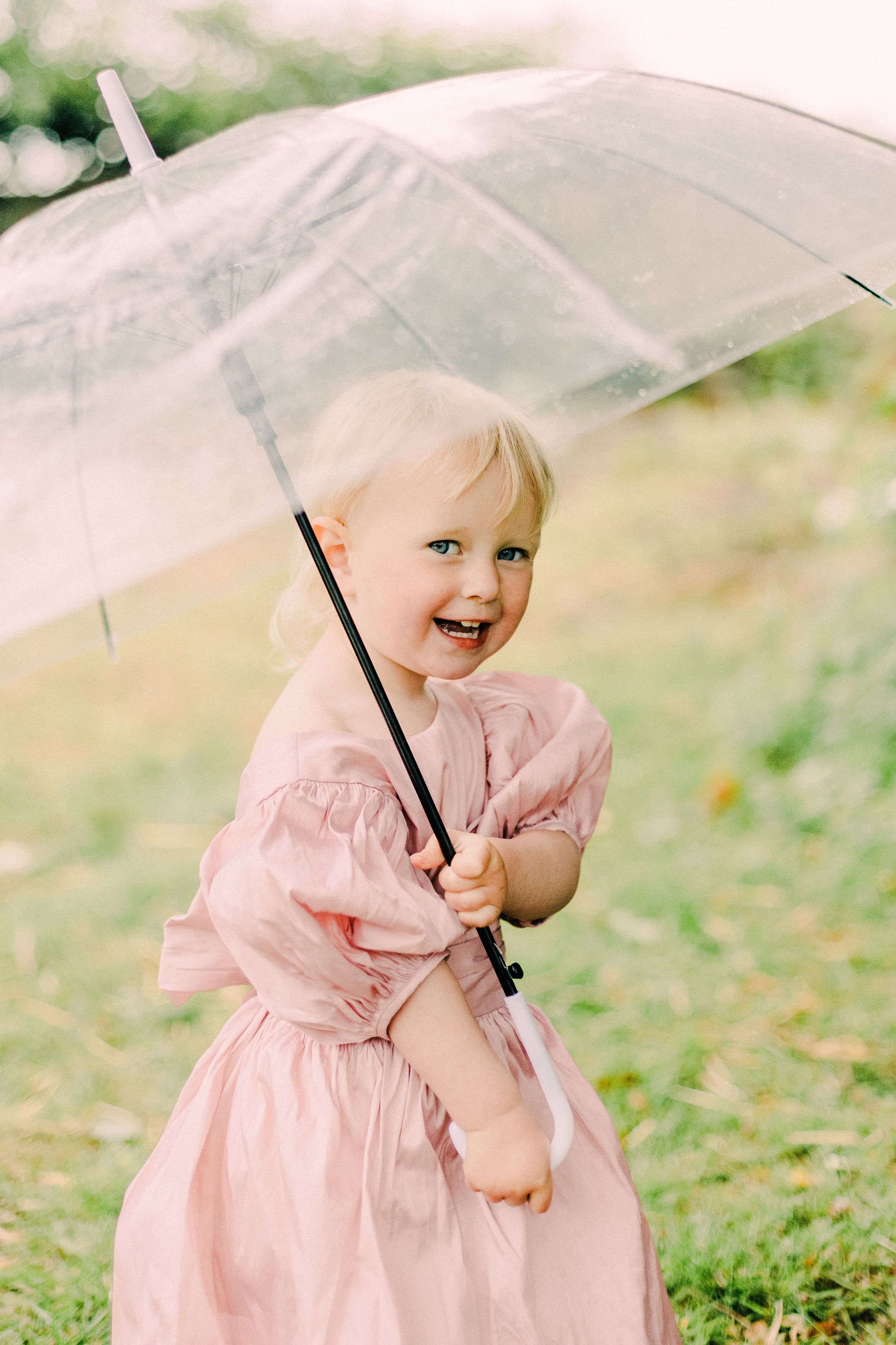 A small child holds a clear umbrella while wearing a pink dress and smiling on a grassy area outdoors.