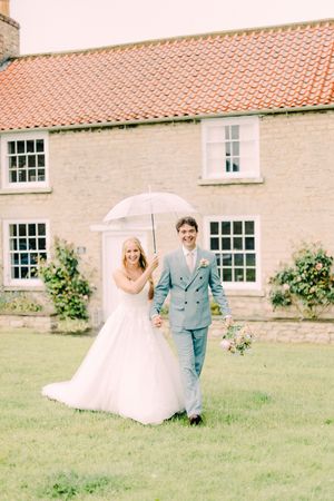 Newlyweds share a romantic moment walking with a white umbrella outside a charming stone cottage with terracotta roof tiles.