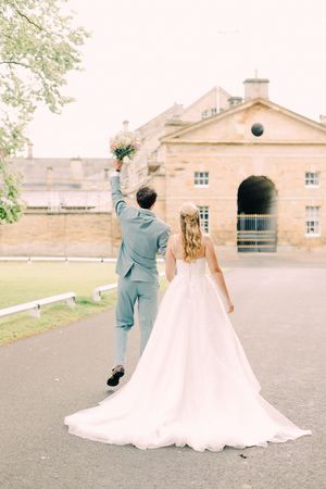 A newlywed couple in pastel blue suit and flowing white gown walking together on a path near a historic building.