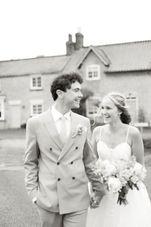 Black and white candid wedding photo of a newlywed couple walking and laughing together by a countryside house.