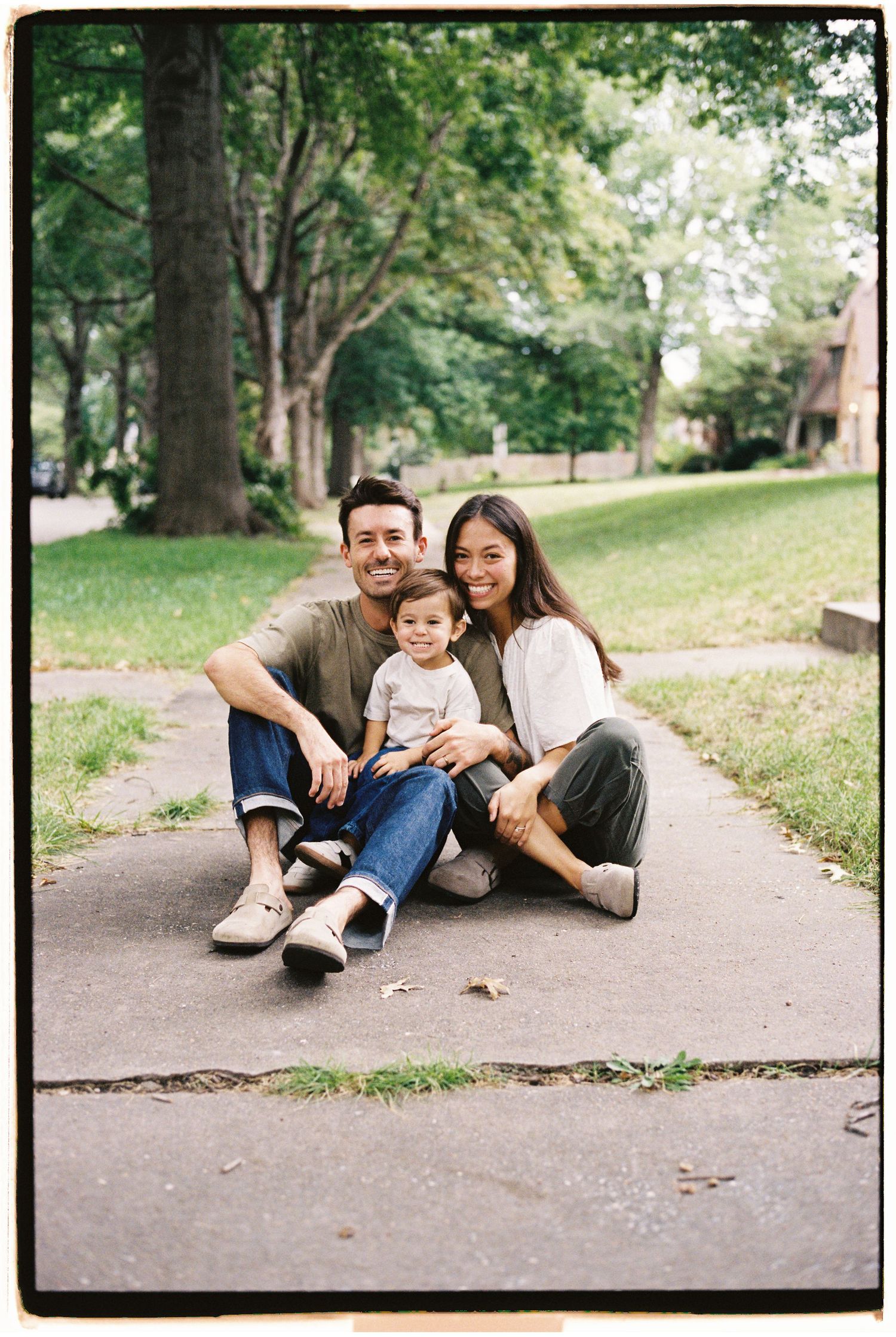 A family relaxes together on a paved pathway surrounded by green grass and trees on a sunny day.