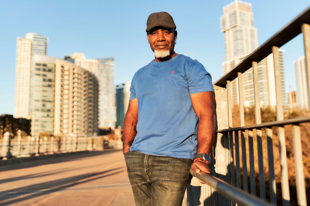 A person in a blue t-shirt and jeans stands on a bridge with city high-rises visible in the warm sunset light.