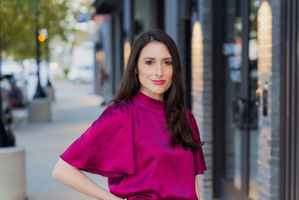 A woman in a bright magenta blouse poses on a city sidewalk with storefronts and trees in the background.