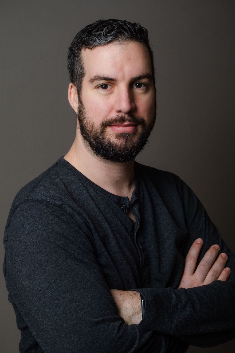 Professional portrait of a person wearing a dark henley shirt against a neutral background with arms crossed.