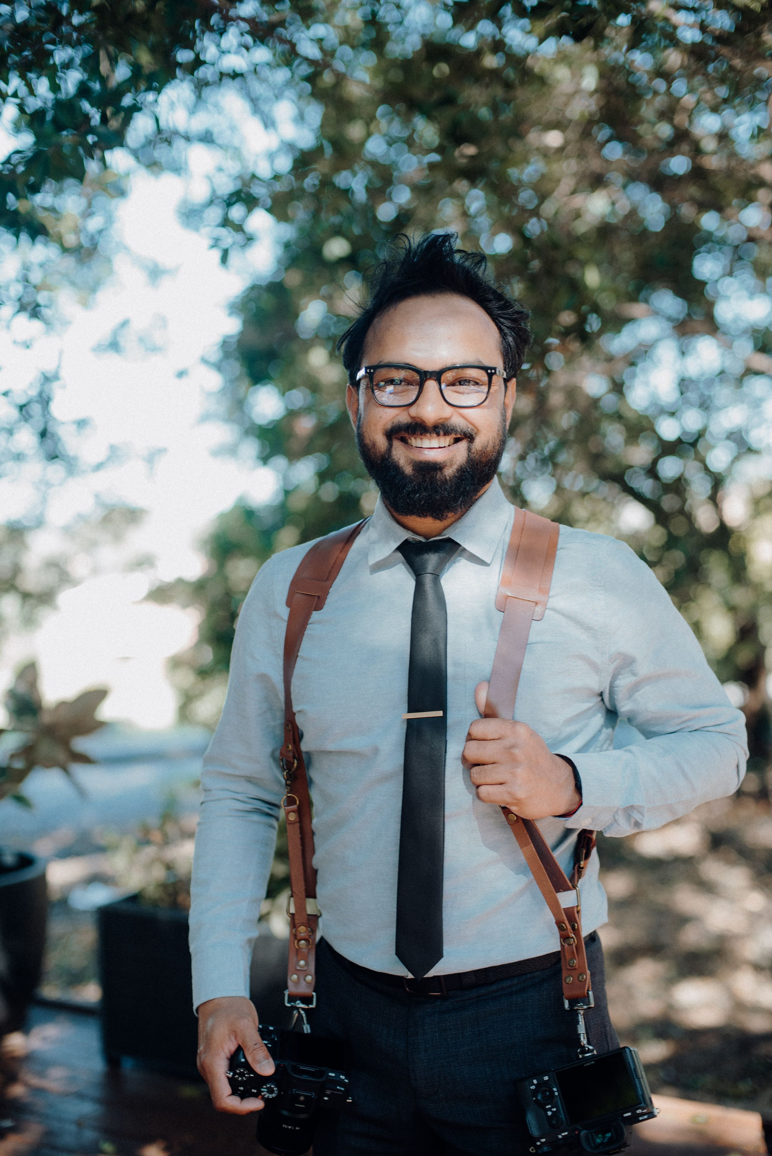 Professional photographer wearing suspenders and glasses smiles while holding cameras outdoors.