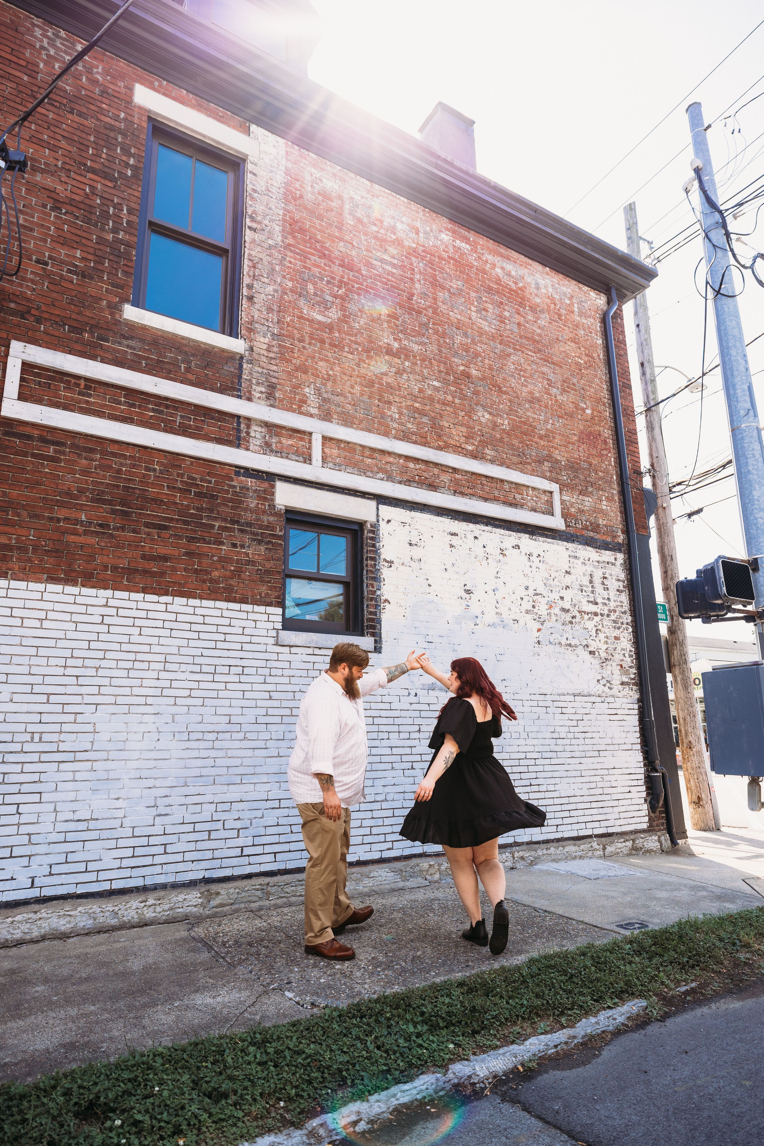 Couple dancing together on a sidewalk beside a rustic brick building in an urban alley on a sunny day.
