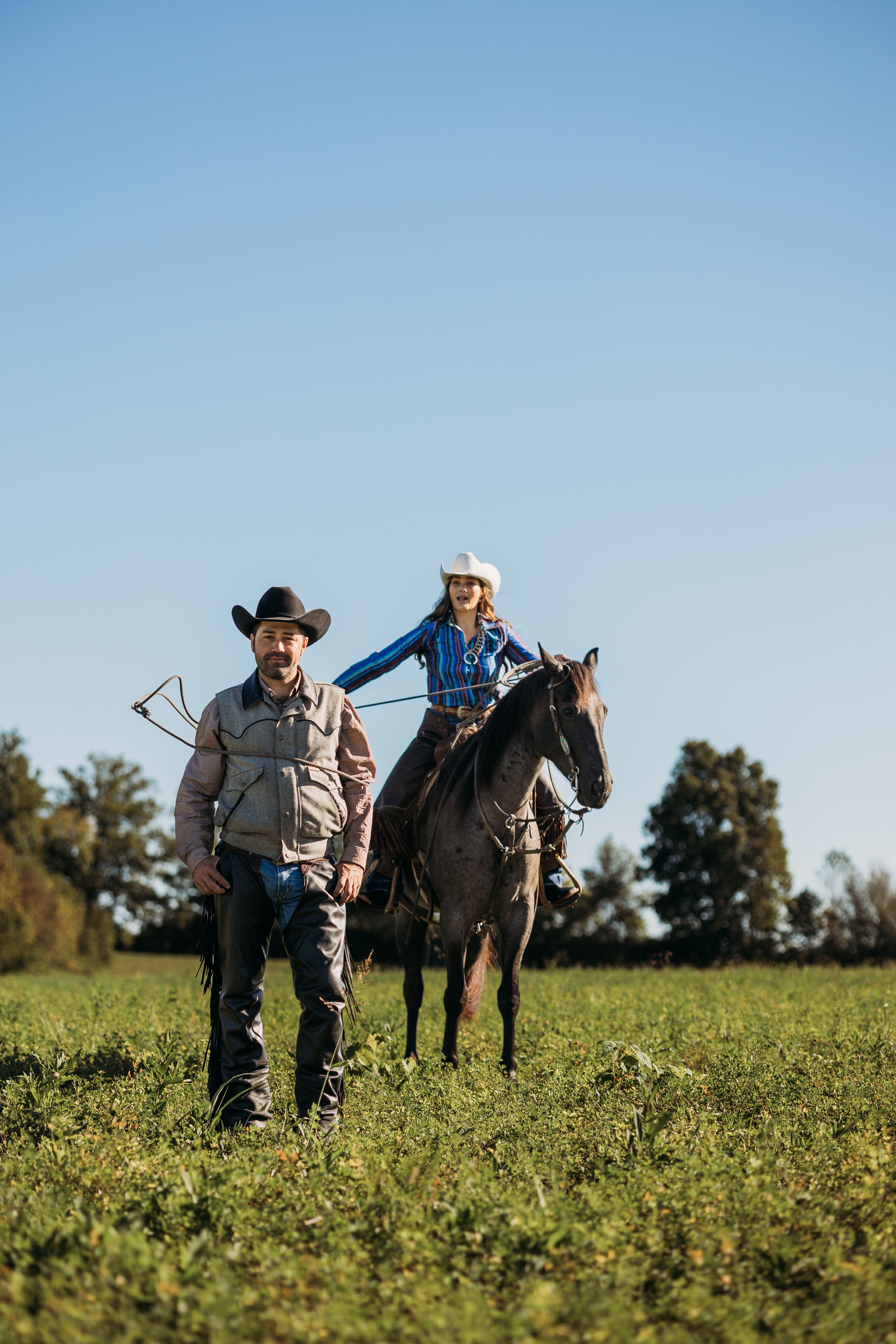 A cowboy walks beside a woman riding a dark horse across a green field under a clear blue sky.