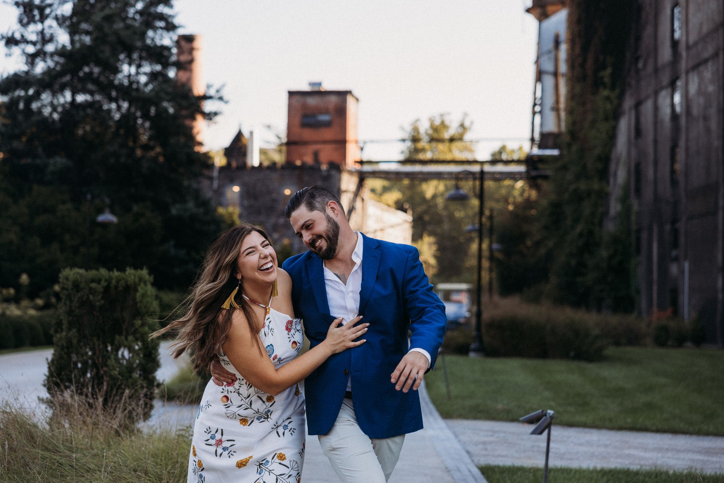 Couple laughing and embracing outdoors at dusk near ivy-covered brick building in elegant wedding attire.
