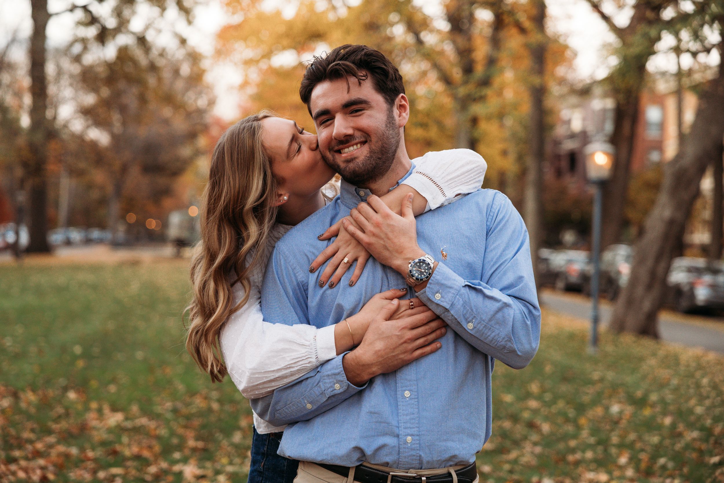 A couple embraces and shares a kiss in an autumn park, with the woman hugging the smiling man from behind.
