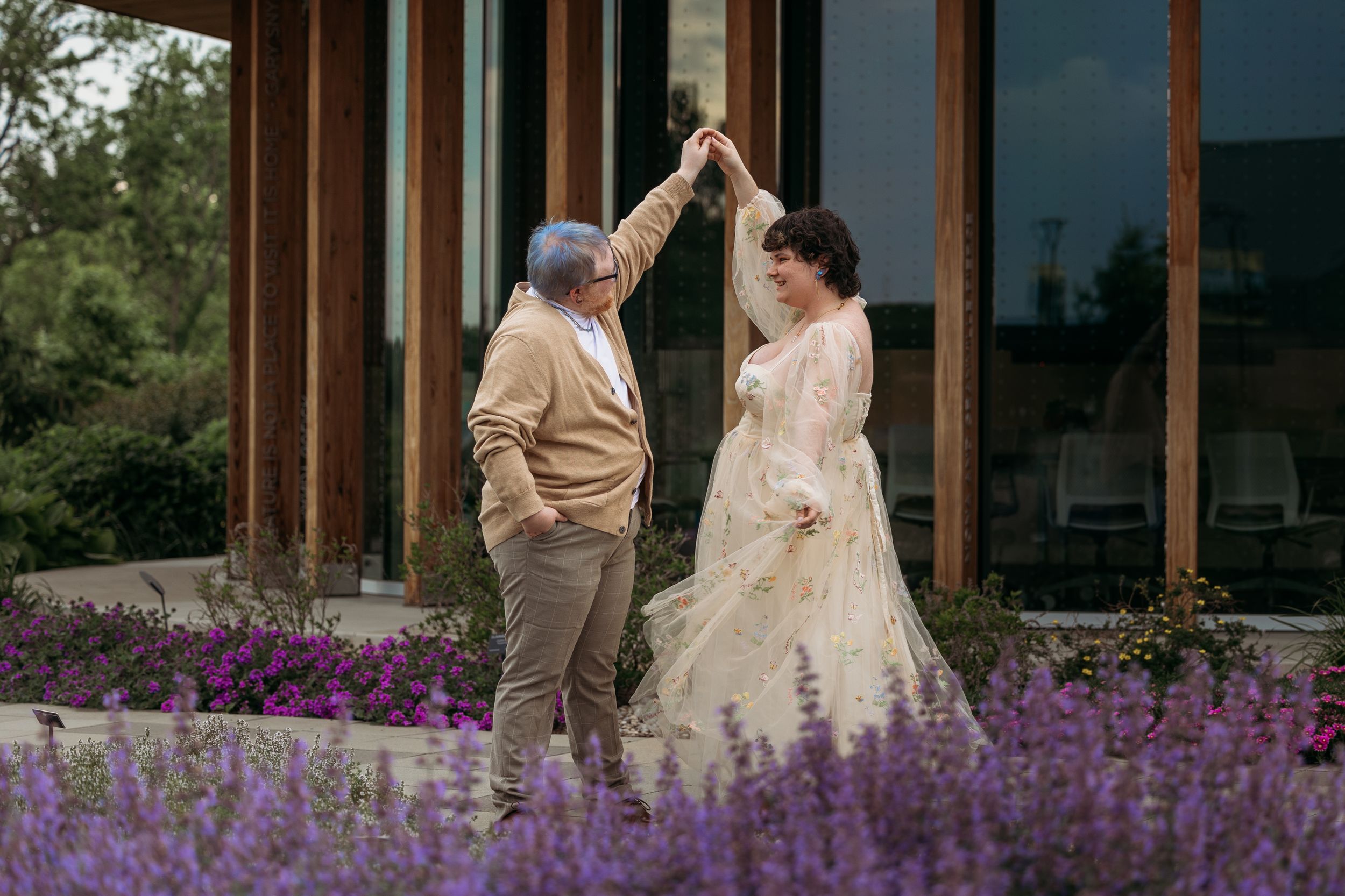 Bride and groom dancing outdoors among purple lavender flowers near a modern building.