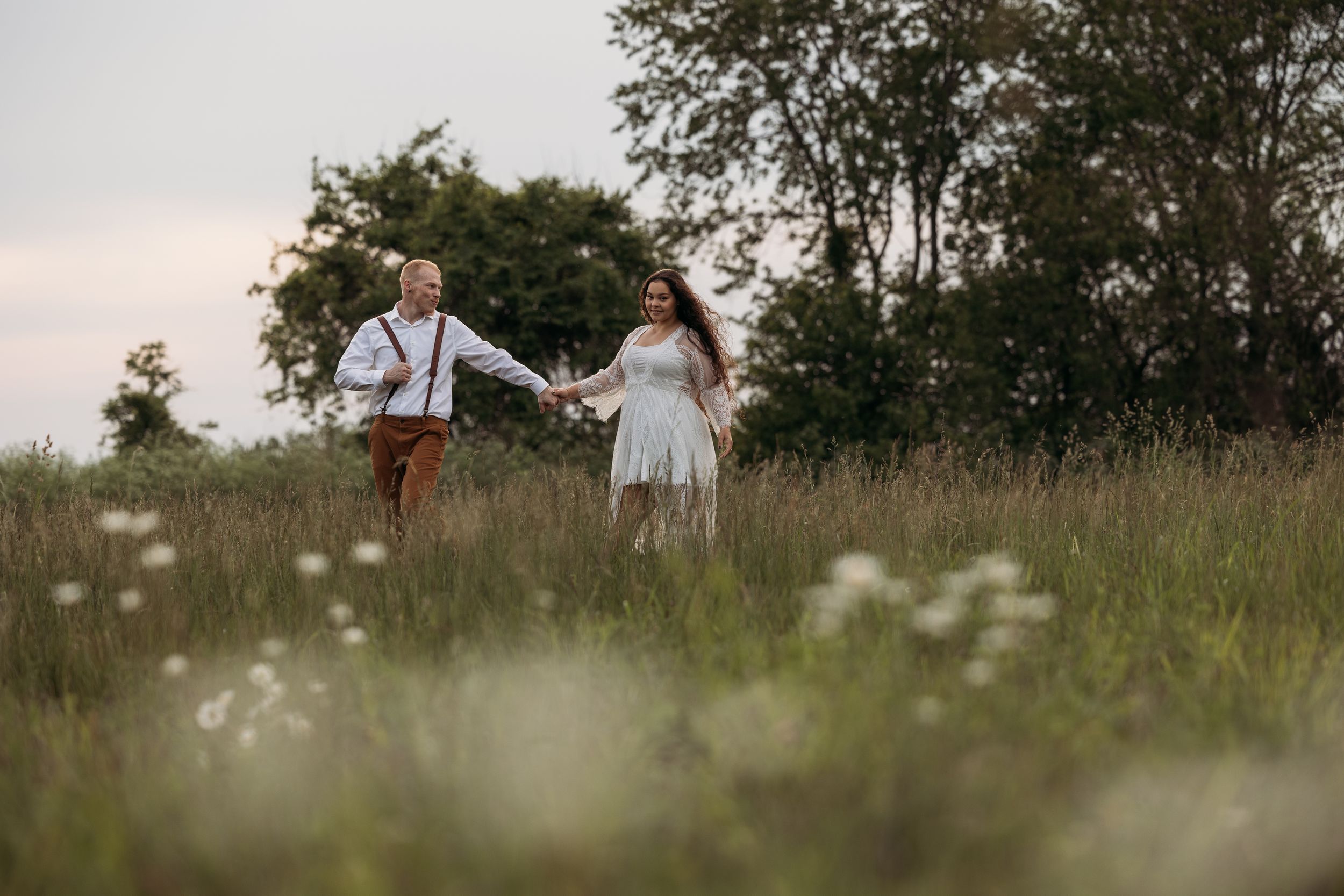Couple holding hands walking through a wildflower meadow with lush green trees in the background at dusk.