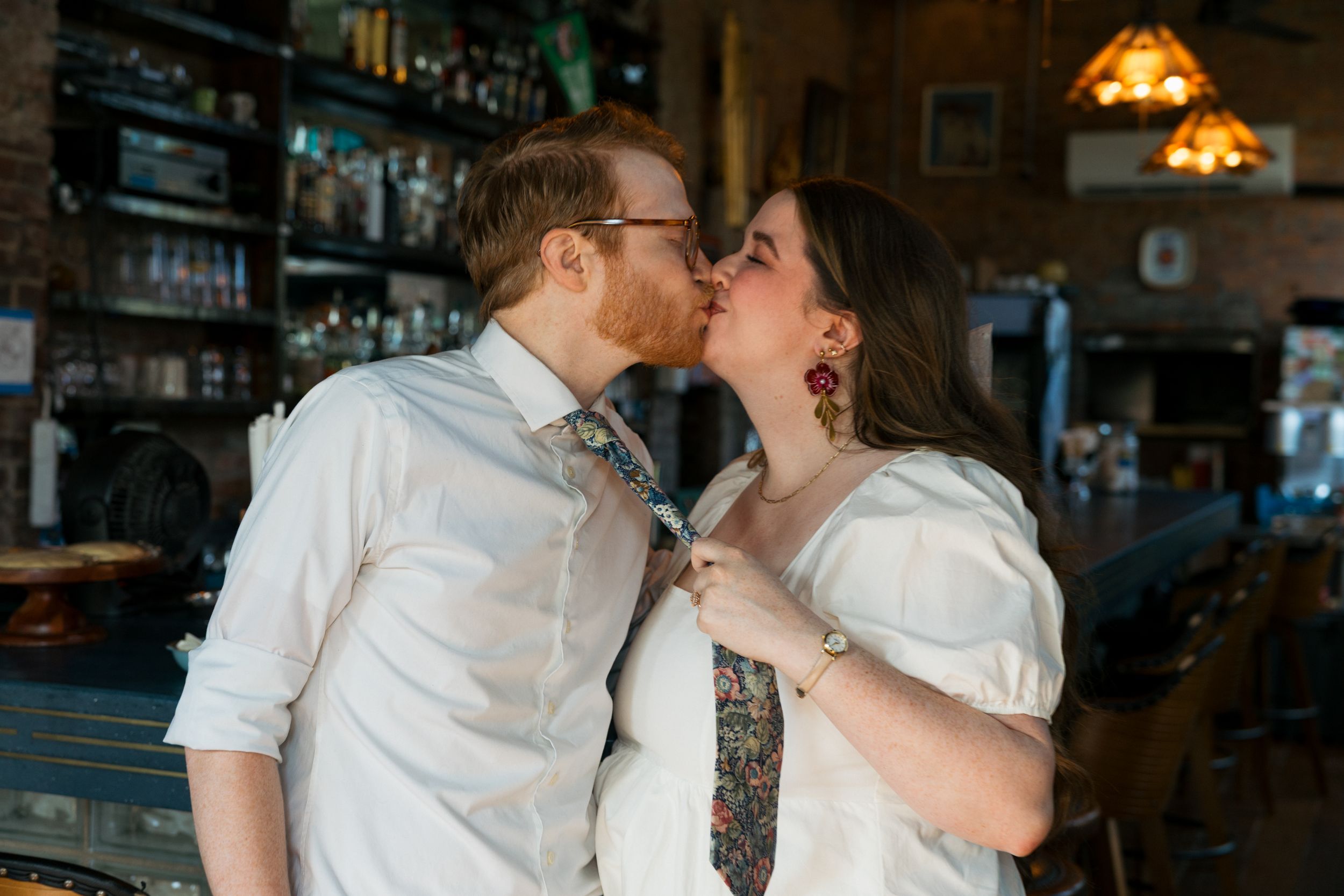 Couple sharing a romantic kiss at a bar, woman holding mans floral tie, both wearing white shirts.