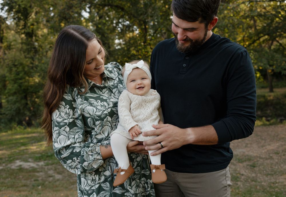 A sweet family moment captured outdoors with a baby in white against a natural backdrop of trees.