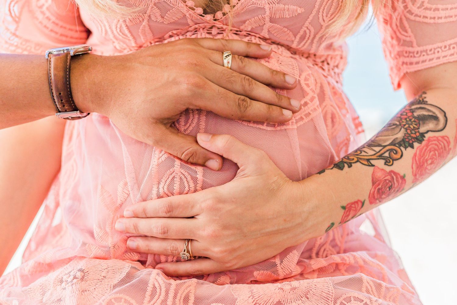 Close-up of hands resting on maternity belly with pink lace dress showing ring and tattoo details at worth ave clock tower in palm beach FL.