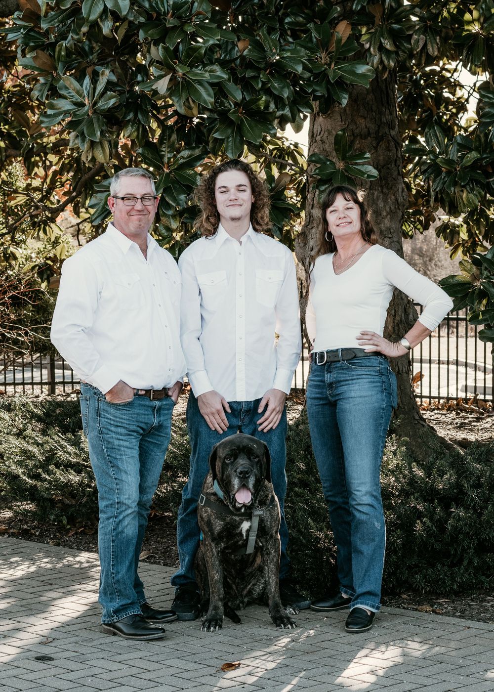Family portrait with three people in white shirts and jeans standing outdoors with their large dog on a stone path.