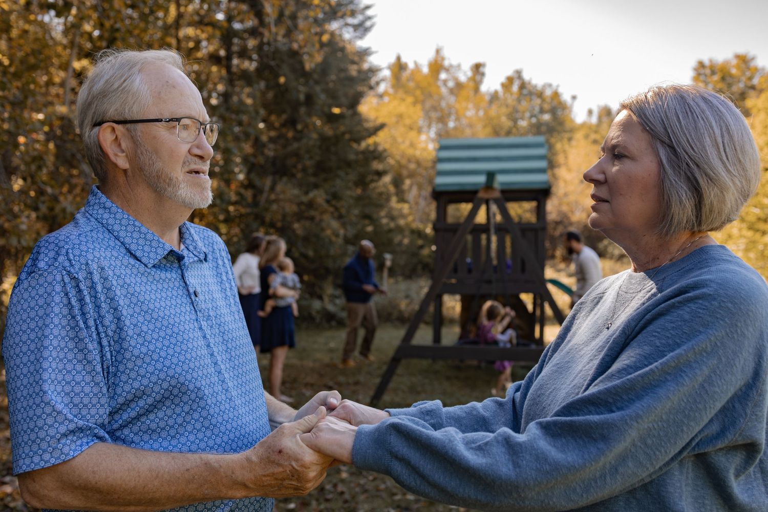 Couple holding hands close together with their children and grandchildren during autumn with golden trees in background.