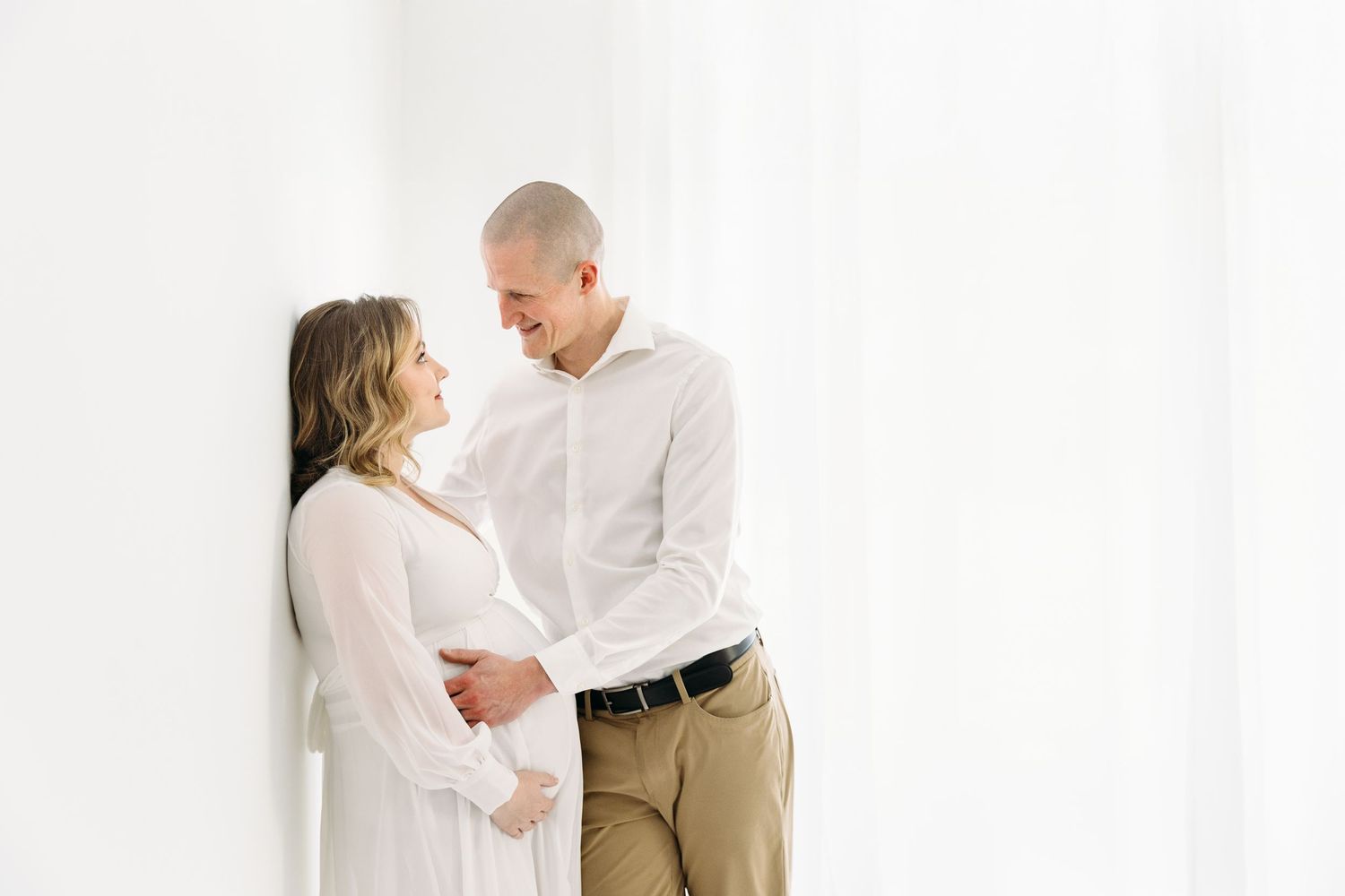 Maternity Photo in a bright white room, featuring a couple in love, looking into each other's eyes, both have their hands on the baby bump.