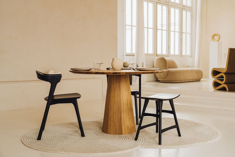 Contemporary dining setup with round wooden pedestal table and black chairs on circular rug.