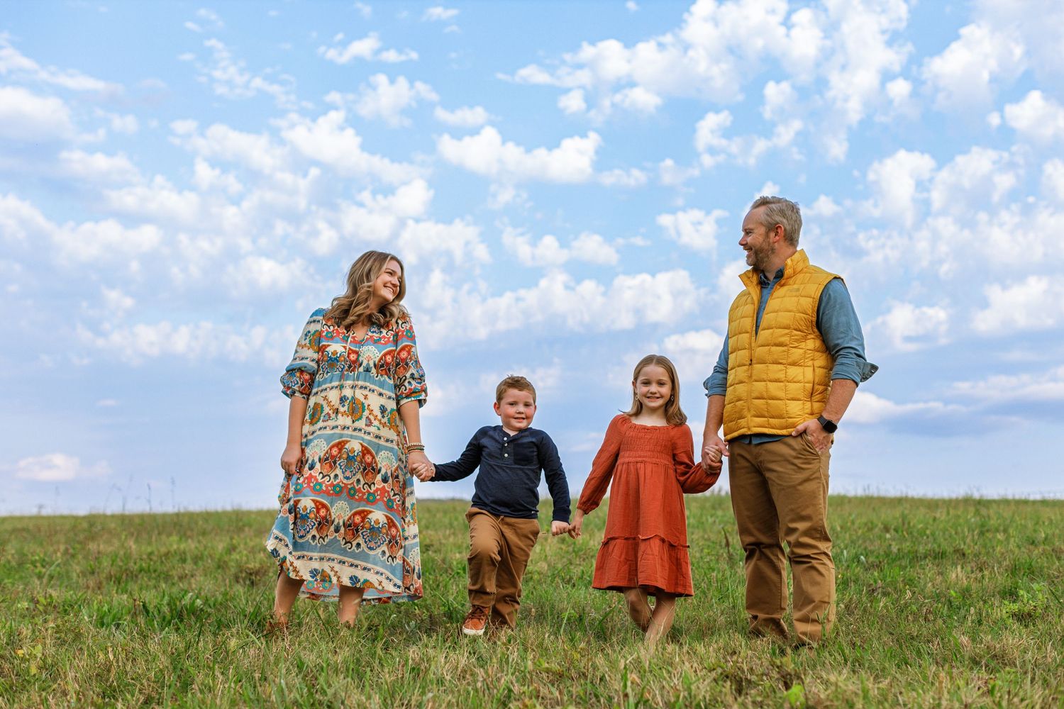 A family of four holds hands while standing together in a green meadow under a bright blue cloudy sky.