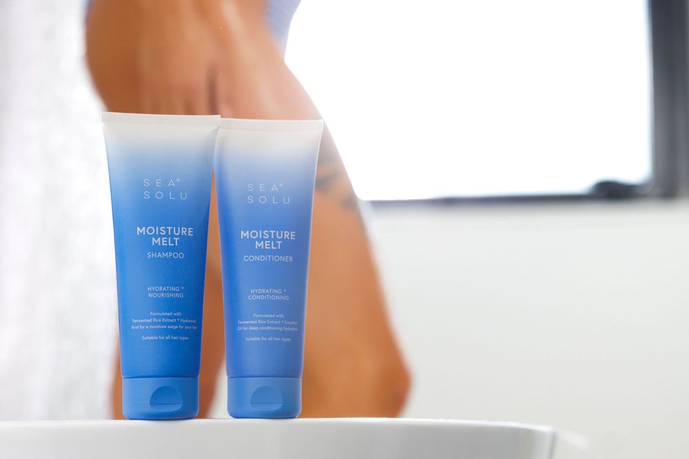 Blue Shampoo & Conditioner tubes arranged minimally on white bathroom surface with someone blurred showering in the background.