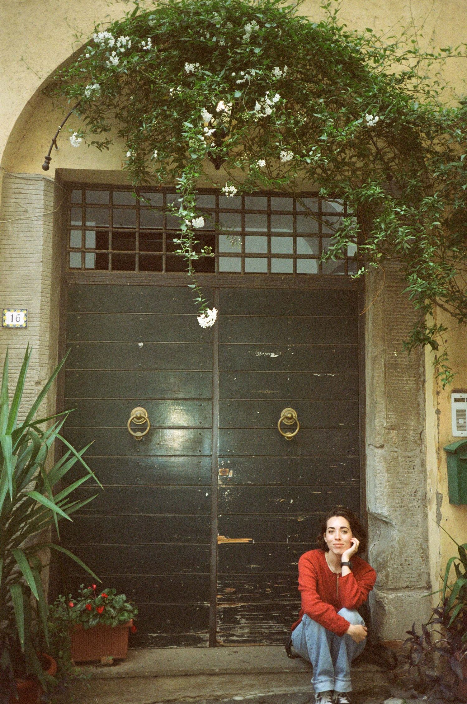 Vintage-style photo of person sitting in front of large black door with climbing plants.