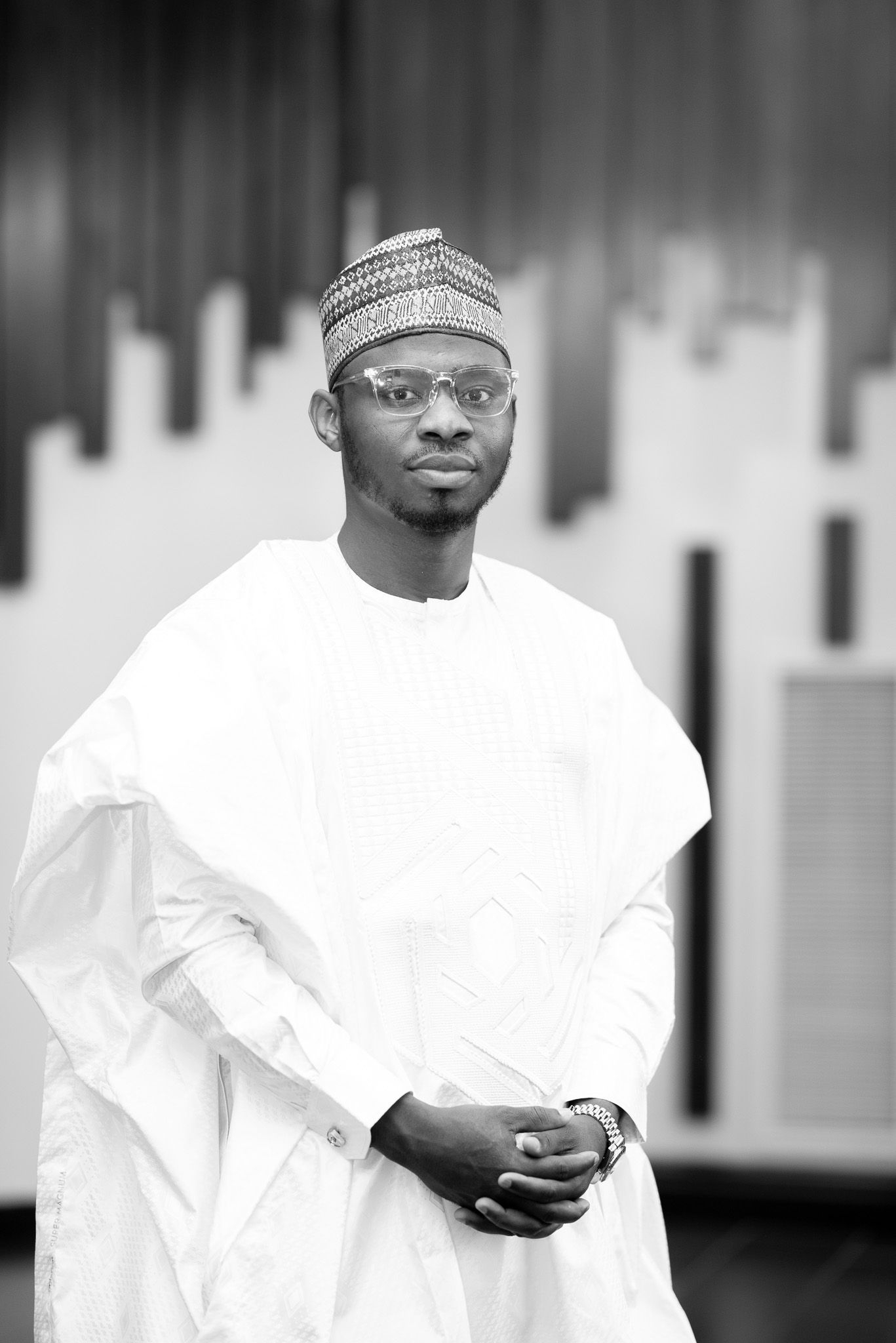 Black and white portrait of a man in traditional West African white attire and kufi cap, posing confidently.