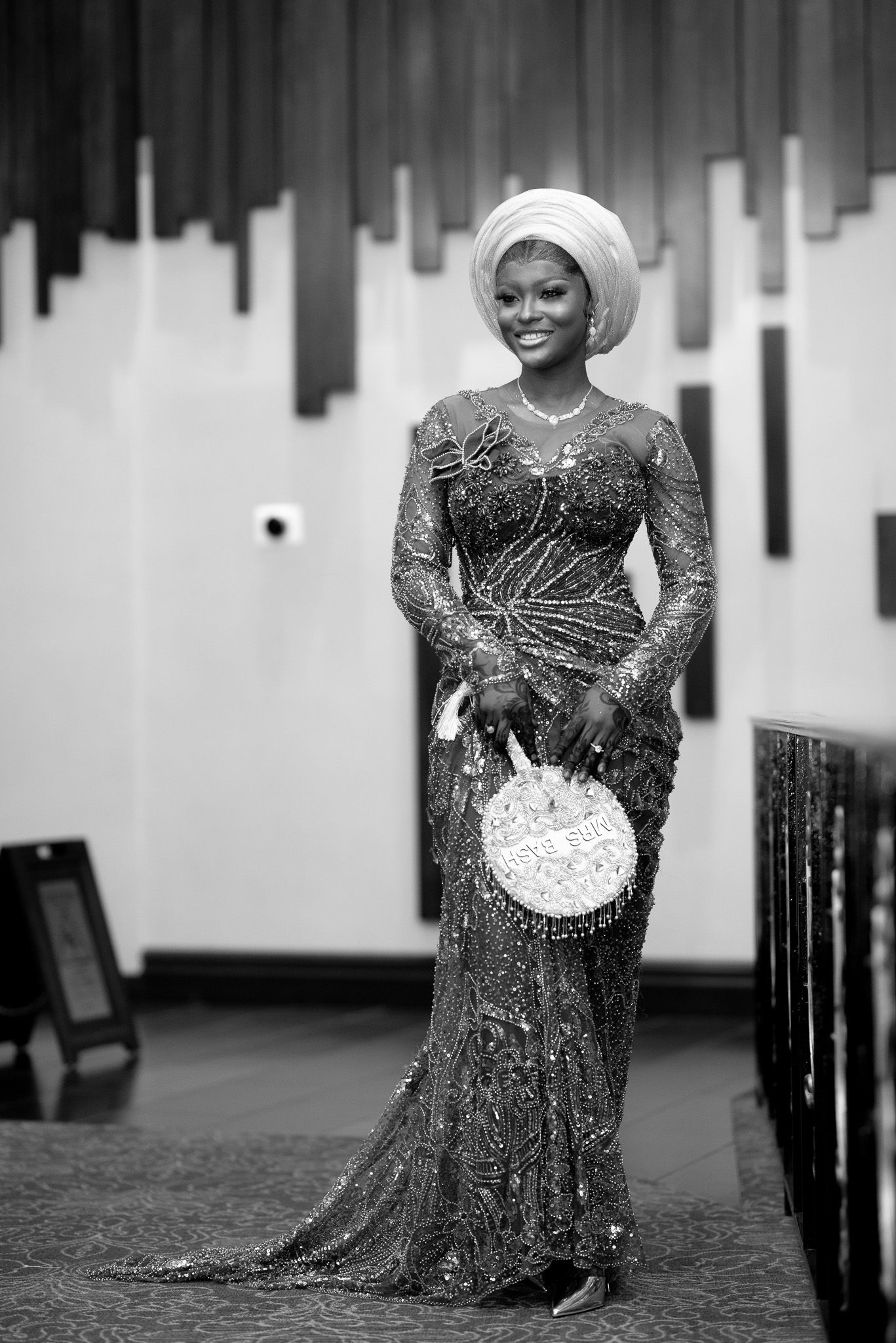 Elegant woman in beaded gown and white gele headwrap poses gracefully in black and white portrait photo.