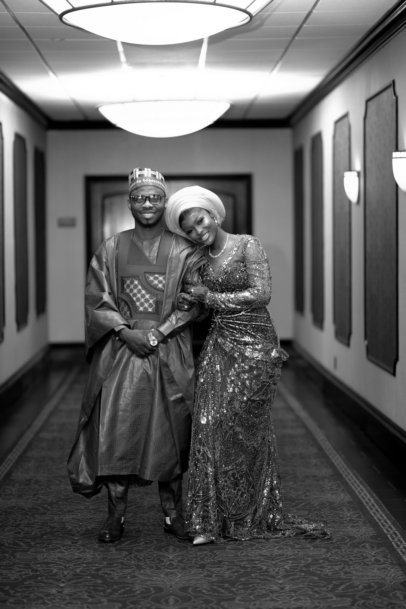 Black and white photo of a couple in traditional African attire posing elegantly in a hallway.