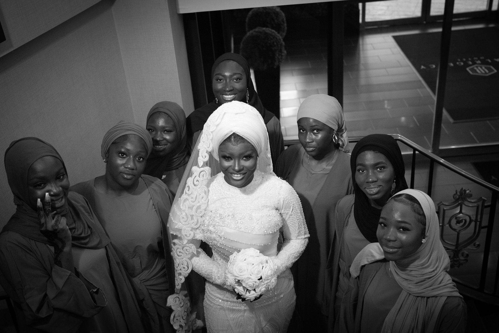 Black and white photo of a bride in white hijab and lace gown surrounded by bridesmaids in hijabs.