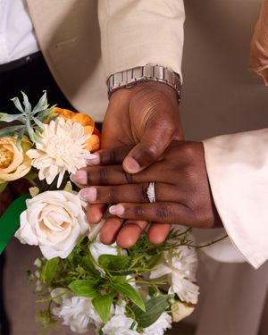 Close-up of hands with wedding rings among white roses and floral arrangements.