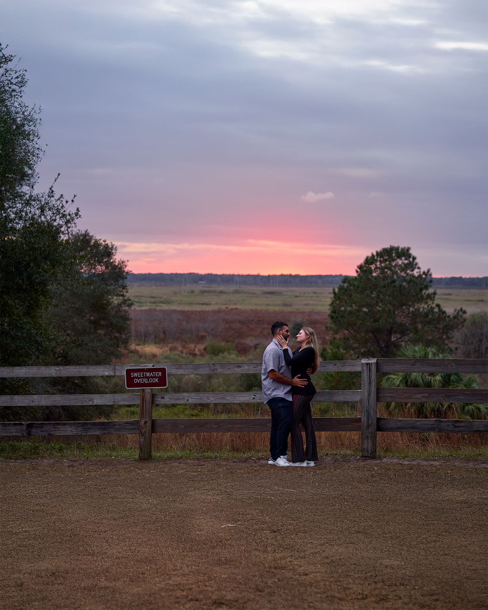 A couple sharing a romantic moment by a wooden fence during a vibrant pink sunset.