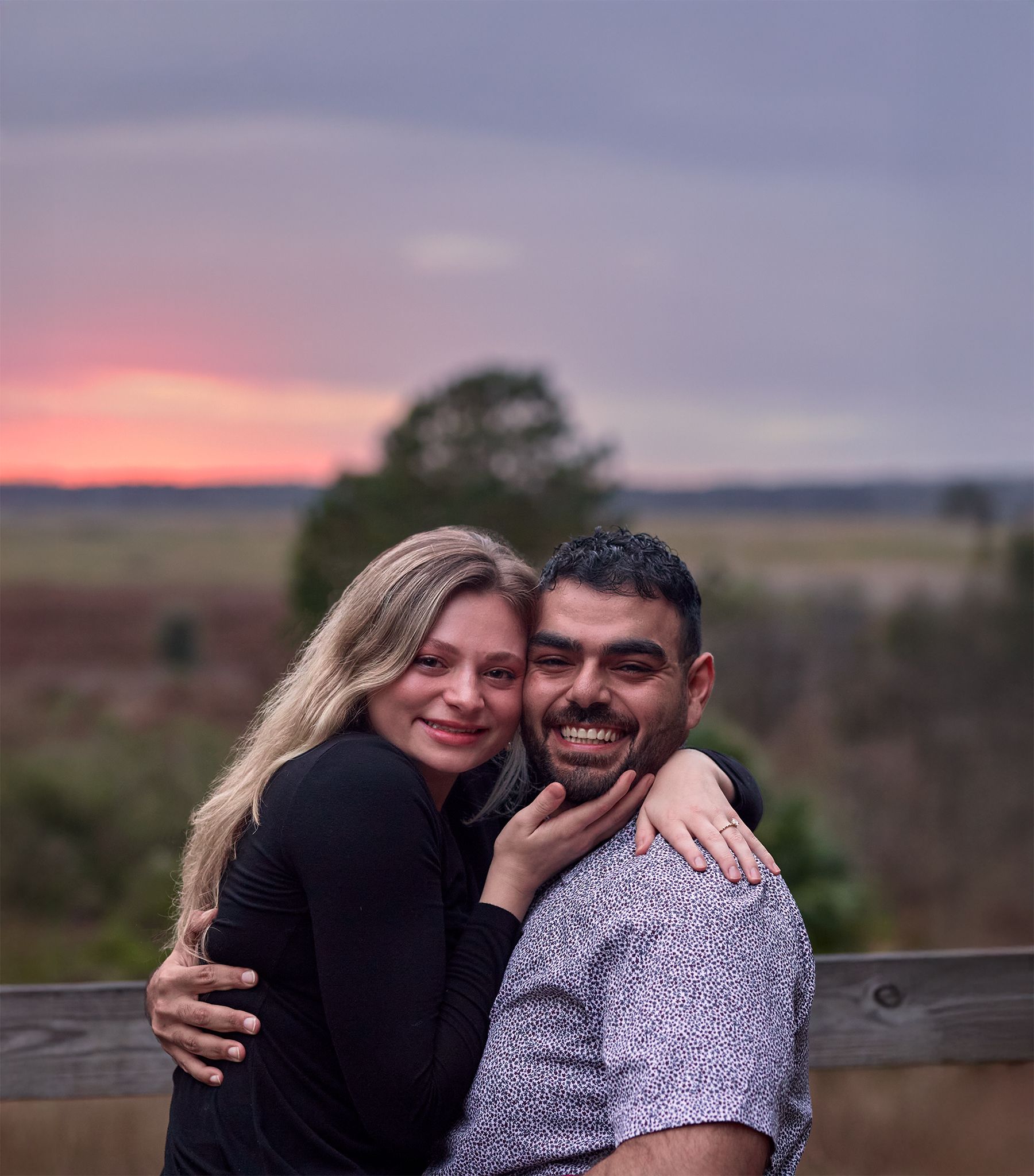 A joyful couple embracing while standing near a wooden fence at sunset.