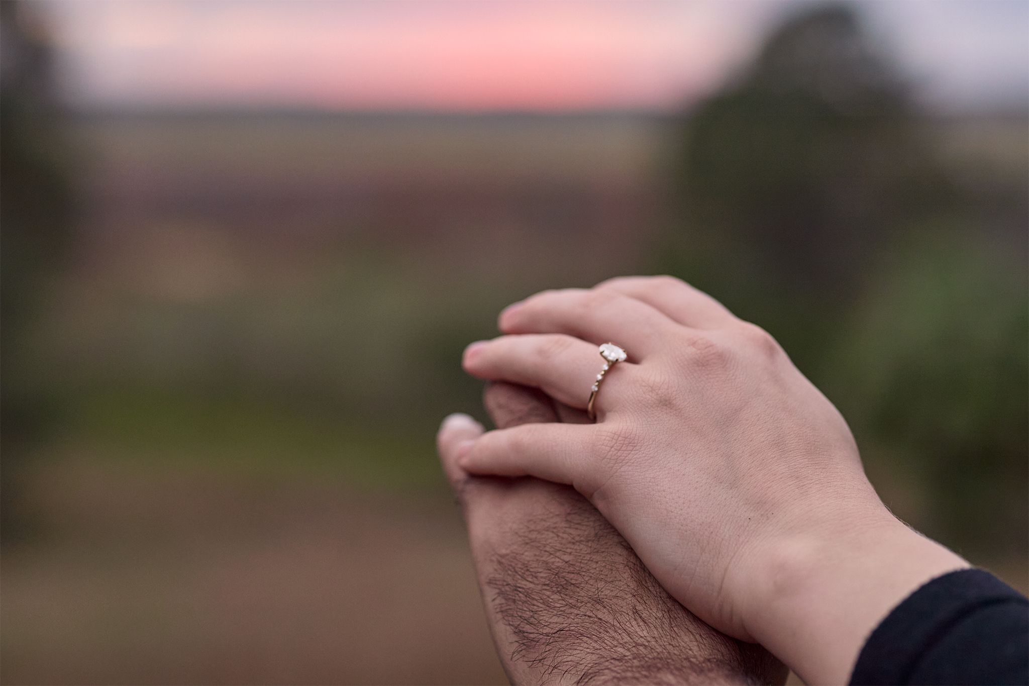 Close-up shot of hands with an engagement ring against a blurred sunset background.
