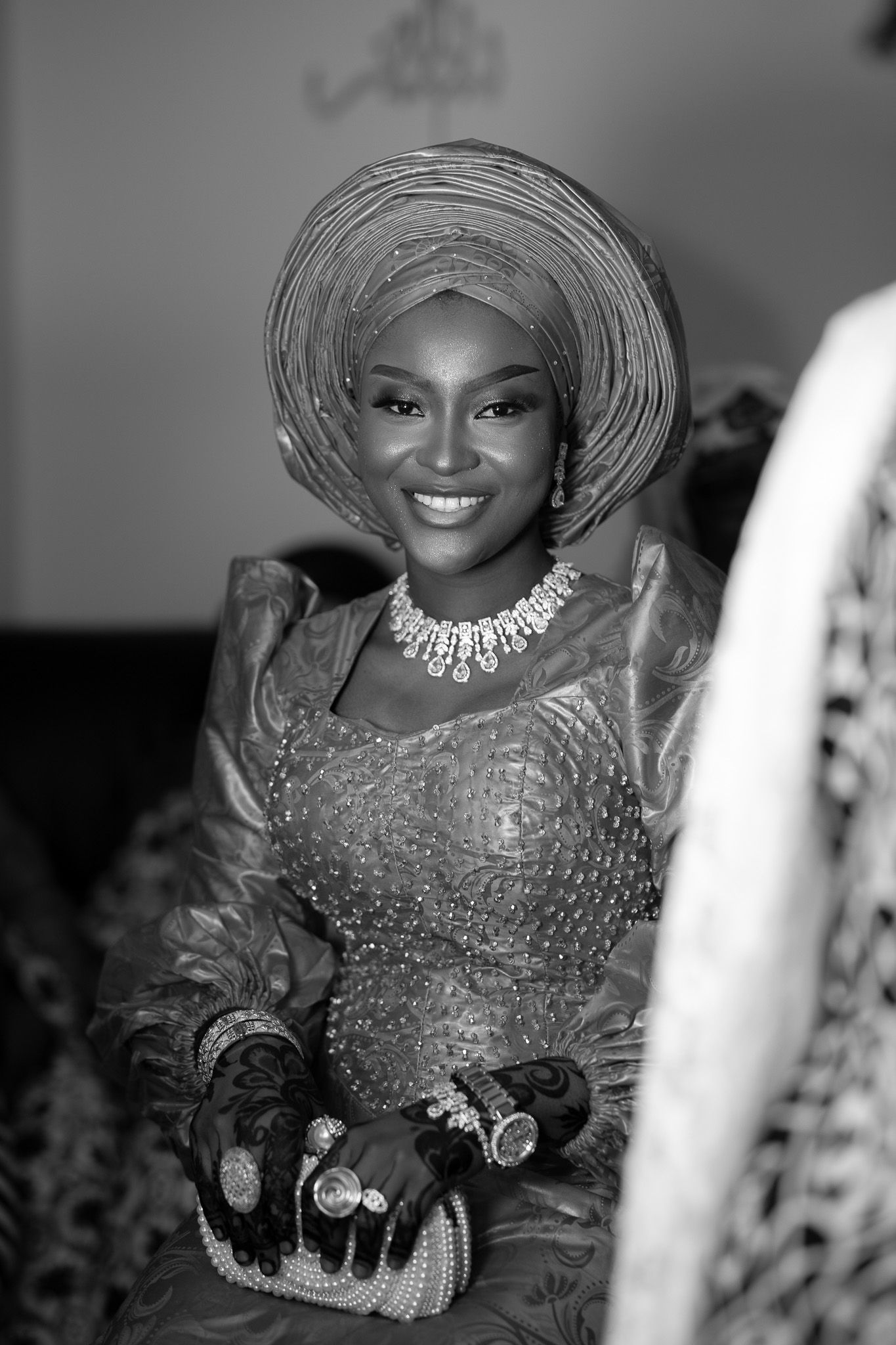 A smiling woman in traditional Nigerian attire, gele headwrap, lace dress, and diamond necklace, captured in black and white.