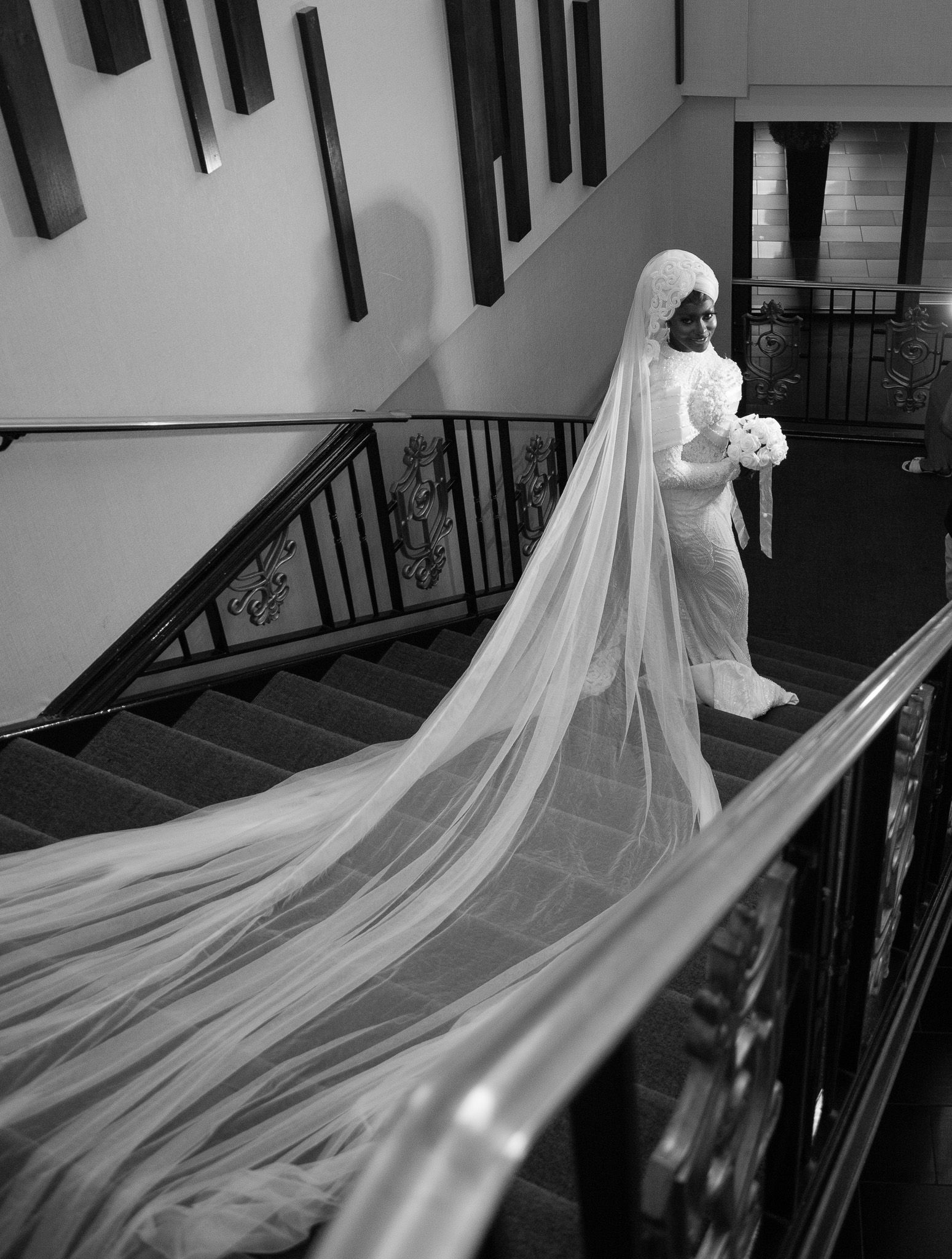Black and white photo of a bride in a long veil descending a grand staircase holding a bouquet.