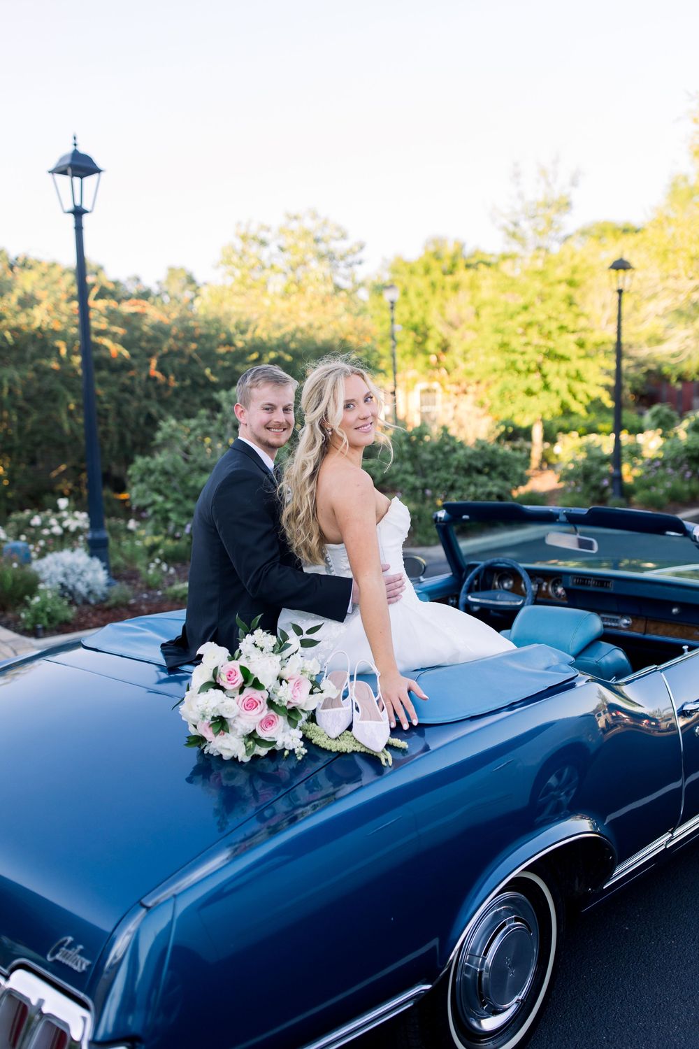 Bride and groom posing on a classic blue convertible car with a floral bouquet, surrounded by lush greenery.