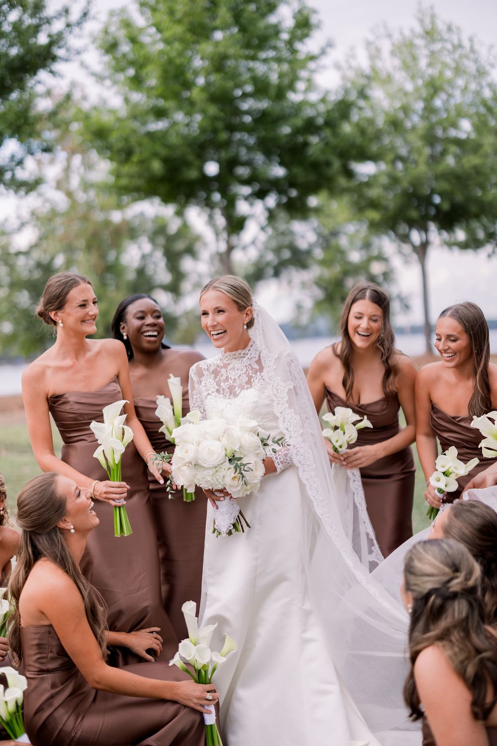 Bridesmaids in brown dresses gather around bride holding white bouquets outdoors.