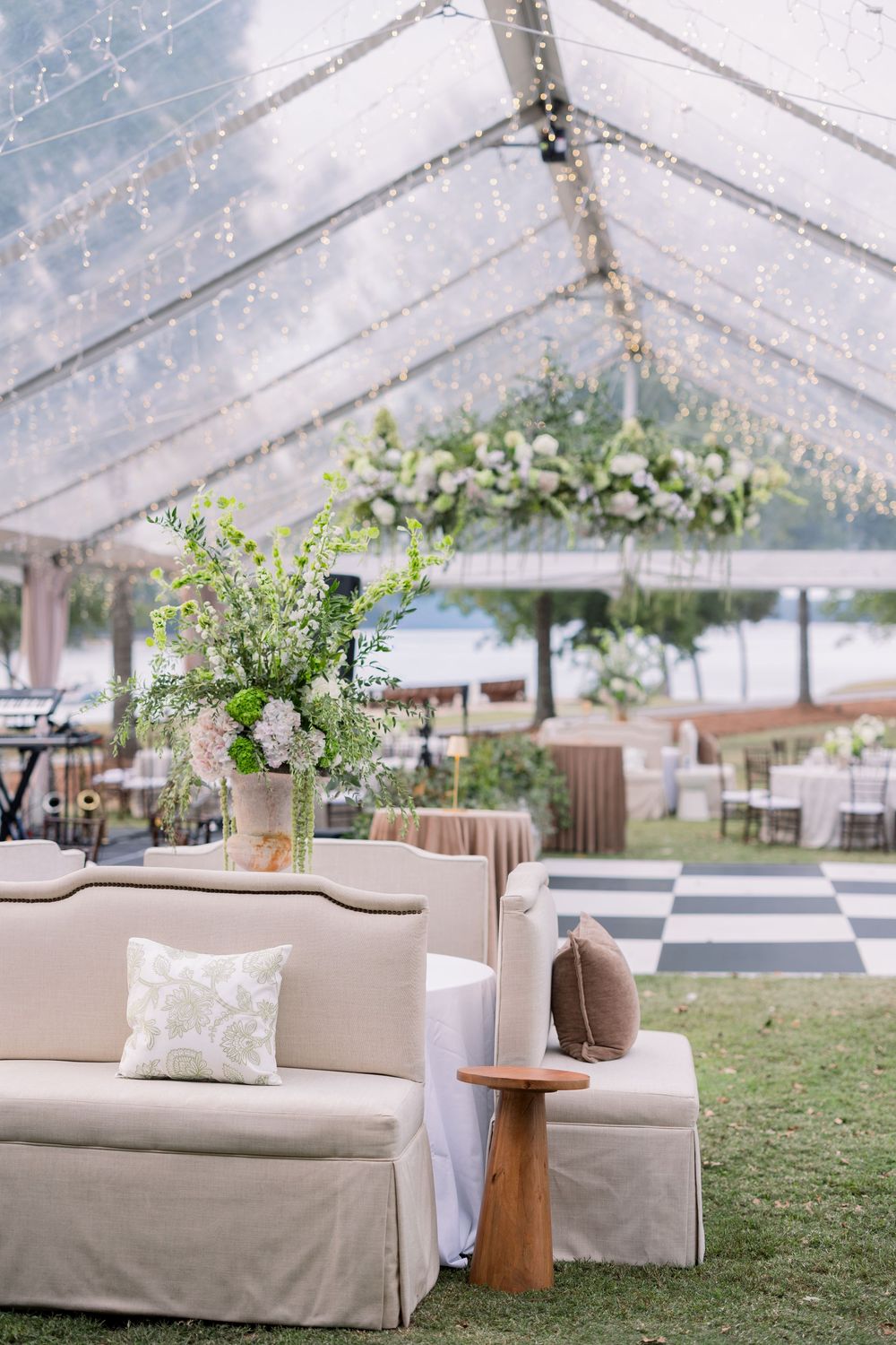 Elegant outdoor wedding lounge area with beige sofa and floral arrangements under clear tent.