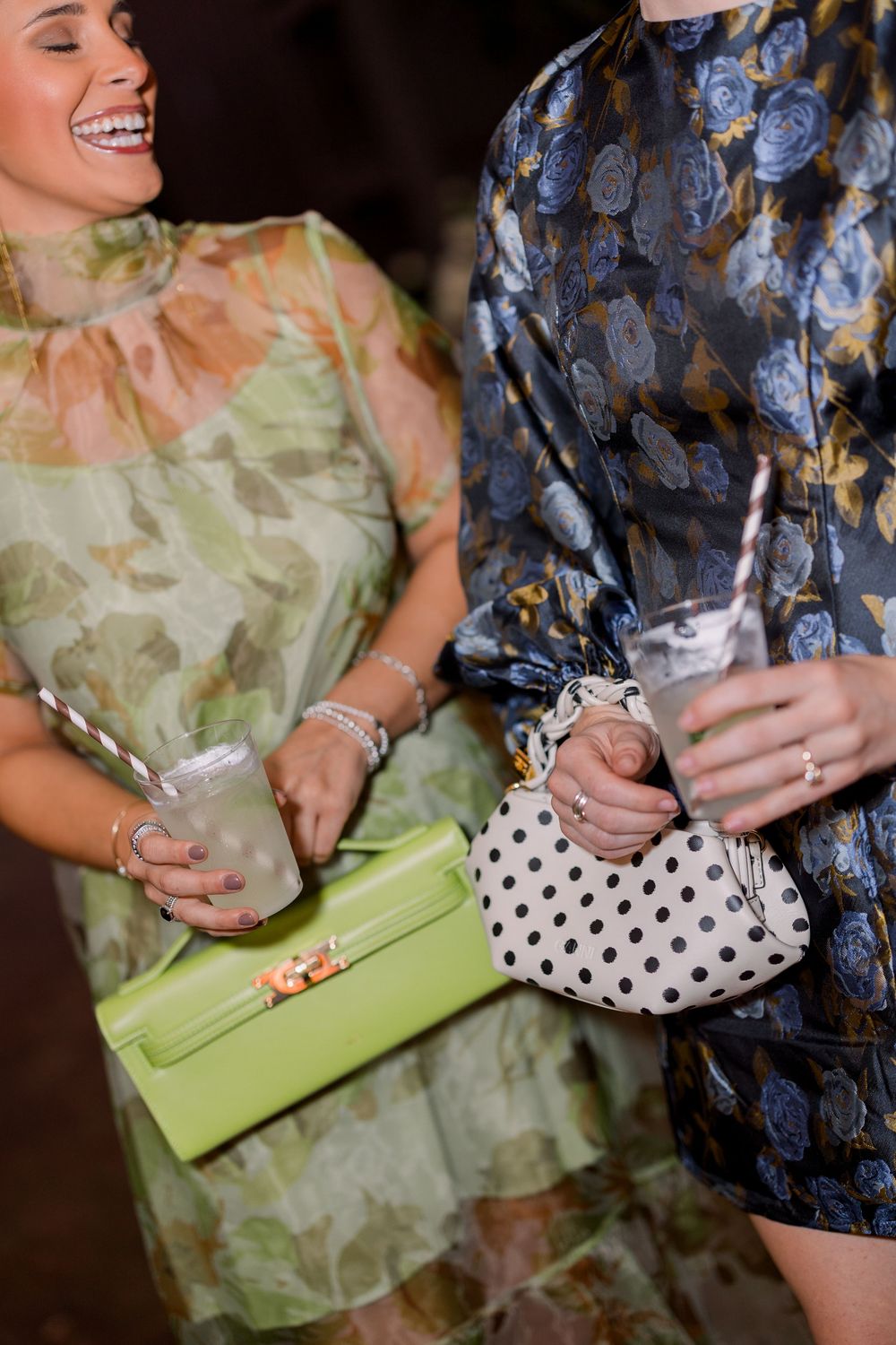 Two people holding drinks and clutches pose together, one wearing a floral dress and one in a polka dot pattern.