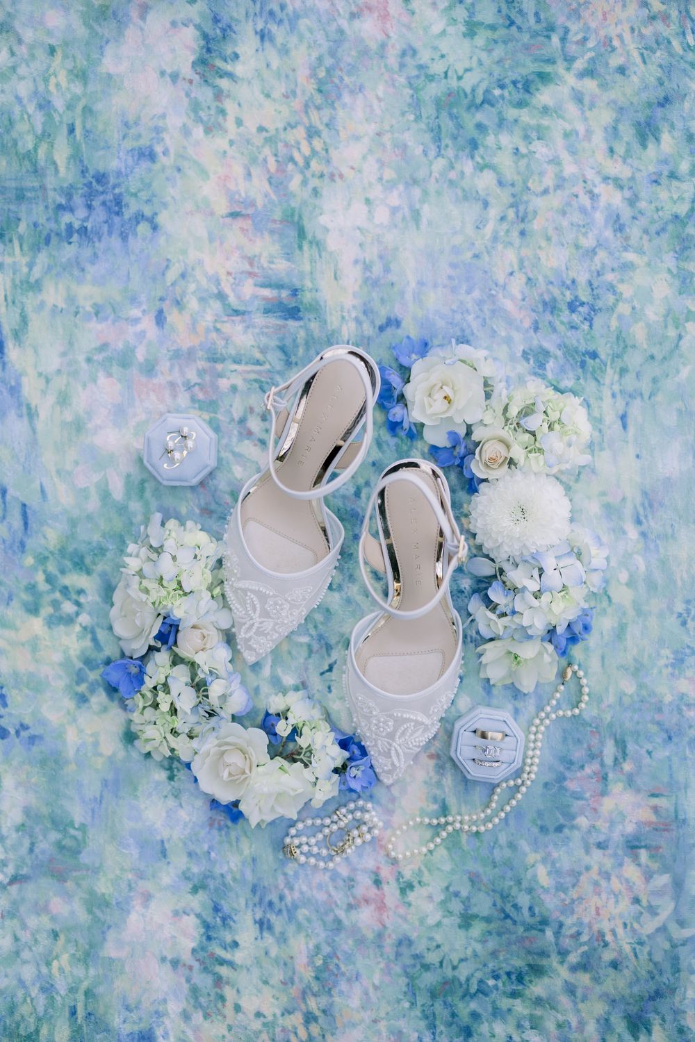 White bridal heels surrounded by white and blue flowers, pearl necklace, and ring boxes on a blue floral backdrop.